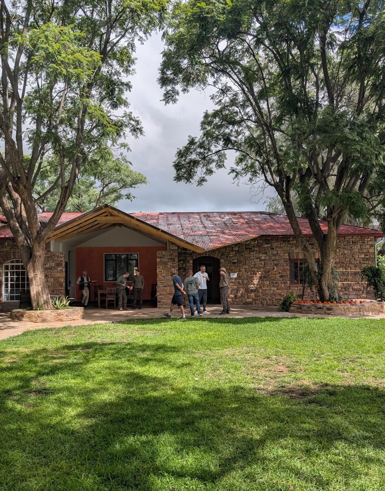 Ein rustikales Steinhaus mit rotem Dach steht zwischen großen Bäumen, während Menschen im Garten plaudern.