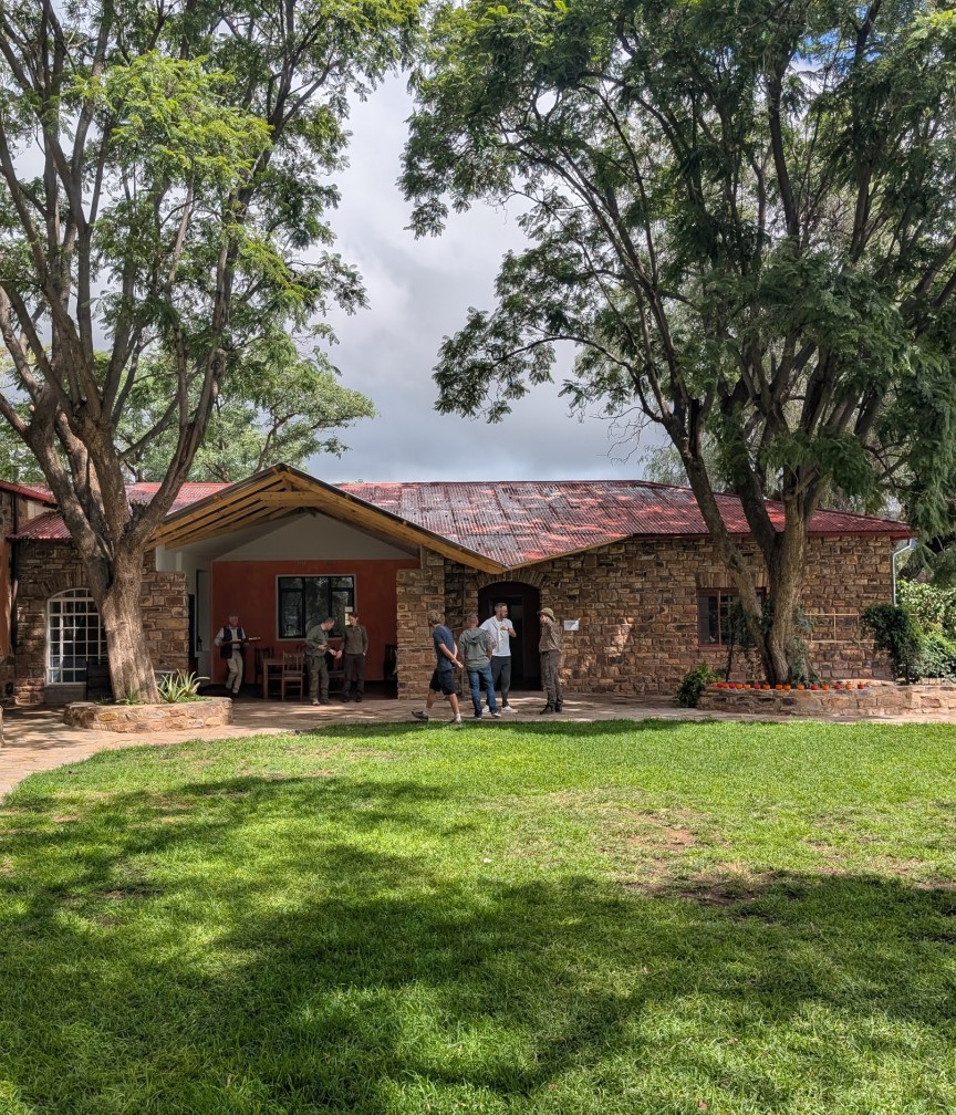 Ein rustikales Steinhaus mit rotem Dach steht zwischen großen Bäumen, während Menschen im Garten plaudern.
